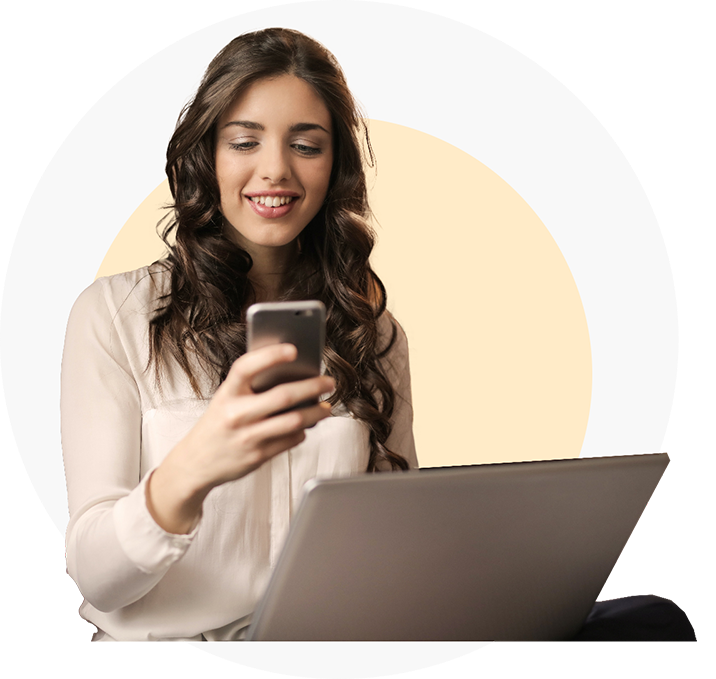 Woman using a smartphone while sitting beside a laptop, focusing on bank reconciliation.