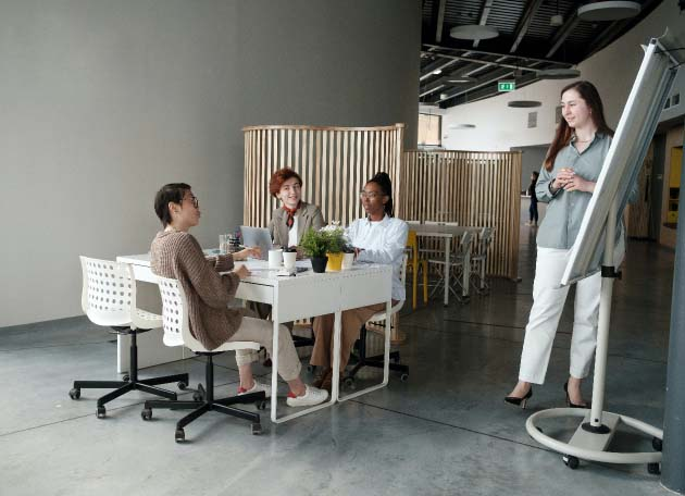 Group of professionals in a modern office setting, engaged in a meeting around a table with a plant, discussing financial and operations management strategies using Dynamics 365 FO. The setting has an open layout with a whiteboard and innovative workspace design.