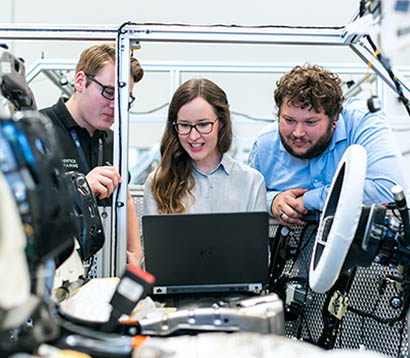 A team of three professionals reviewing data on a laptop in an automotive workshop, illustrating collaboration and technology in financial and operations management with Dynamics 365 FO.
