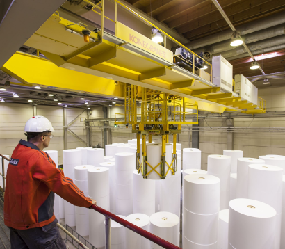 Worker operating a crane to lift large rolls of paper in a manufacturing facility.