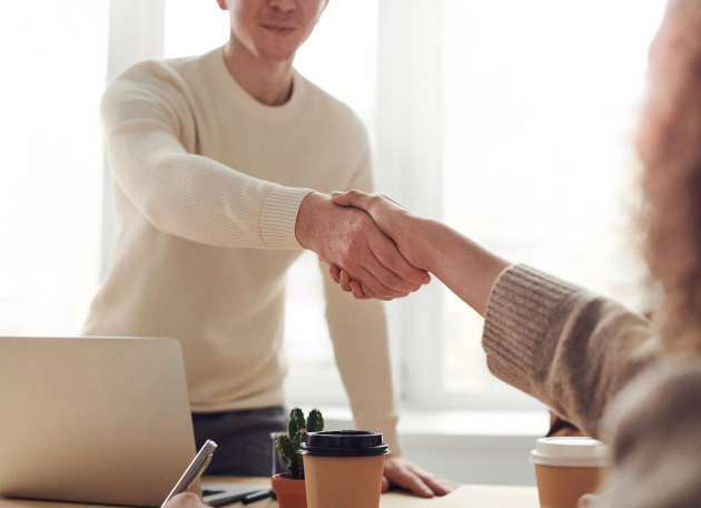 Two people shaking hands in an office environment with a laptop and coffee cups on the table, representing business collaboration and custom software development services by TechnicaX.